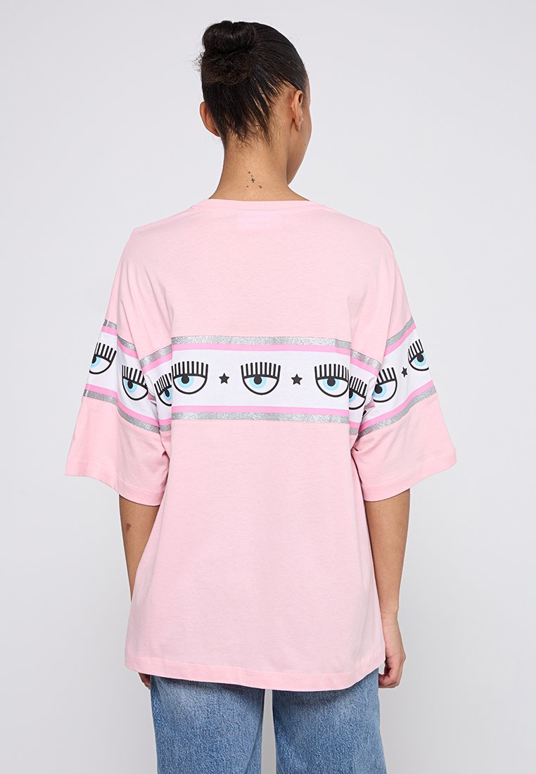 T-shirt rose surdimensionné en coton avec des manches courtes. Présente une bande blanche avec des graphiques d'yeux noirs et bleus et des rayures argentées.