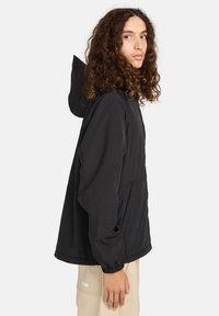 Junge Person mit langen lockigen Haaren, die eine schwarze Kapuzenjacke und beige Hosen trägt, steht seitlich vor einem einfarbigen Hintergrund.