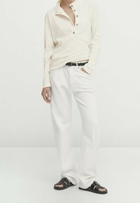 Krämfärgad, långärmad henley-tröja med knapprad, kombinerad med vita, löst sittande byxor och svarta sandaler, vilket visar en avslappnad passform.