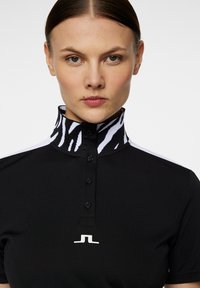 Sort polo T-shirt med zebrastribet krave, fire knapper og hvide accentstriber på skuldrene. Logo vist på brystet.