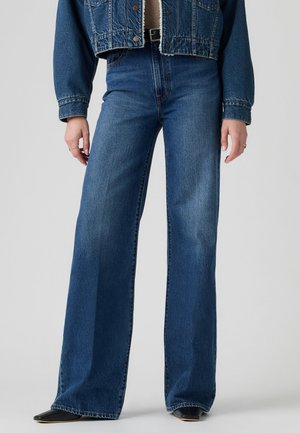 Personne portant un jean bleu taille haute à jambes larges, une veste en jean courte et des chaussures noires pointues, debout devant un fond uni.