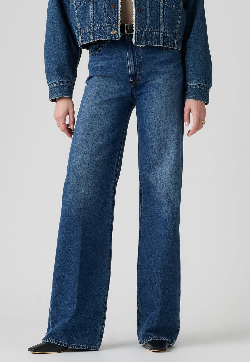 Personne portant un jean bleu taille haute à jambes larges, une veste en jean courte et des chaussures noires pointues, debout devant un fond uni.