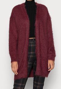 Burgundy fluffig cardigan med öppen front, långa ärmar och textilmaterial. Bärs över en svart ribbad topp och rutiga byxor.