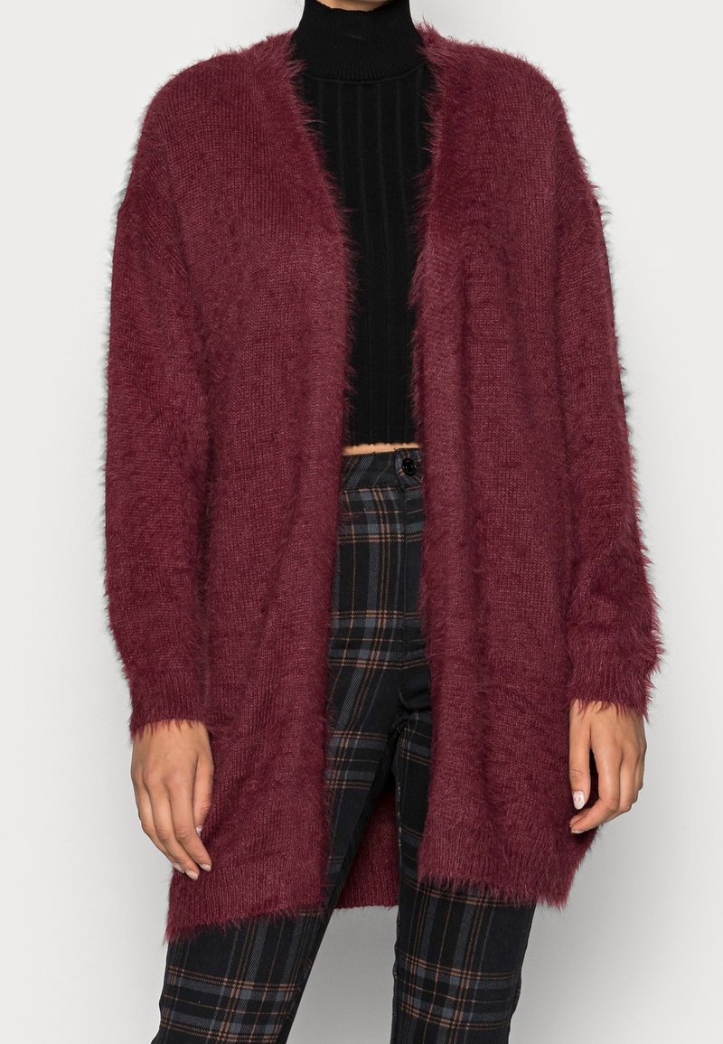 Burgundy fluffig cardigan med öppen front, långa ärmar och textilmaterial. Bärs över en svart ribbad topp och rutiga byxor.