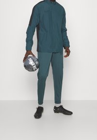 Man in een teal Nike trainingspak die een zwarte en grijze Nike FC voetbal vasthoudt, gekleed in zwarte voetbalschoenen met grijze sokken tegen een witte achtergrond.