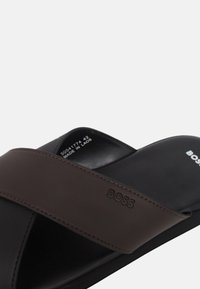 Brązowe i czarne klapki z szerokim nakładającym się paskiem, teksturowanym materiałem oraz wytłoczonym logo marki na wkładce.