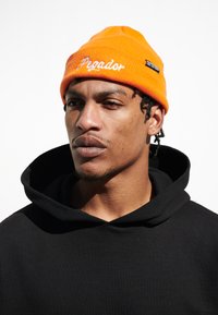 Orange strikbeanie med hvid broderet tekst "Pegar" og et lille sort mærke. Parret med en sort hættetrøje.
