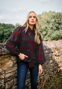 Czarna i burgundowa blouse w kwiaty z bufiastymi rękawami, zapinana na guziki z delikatnymi akcentami w formie ćwieków. Noszona na ciemnoniebieskich dżinsach.