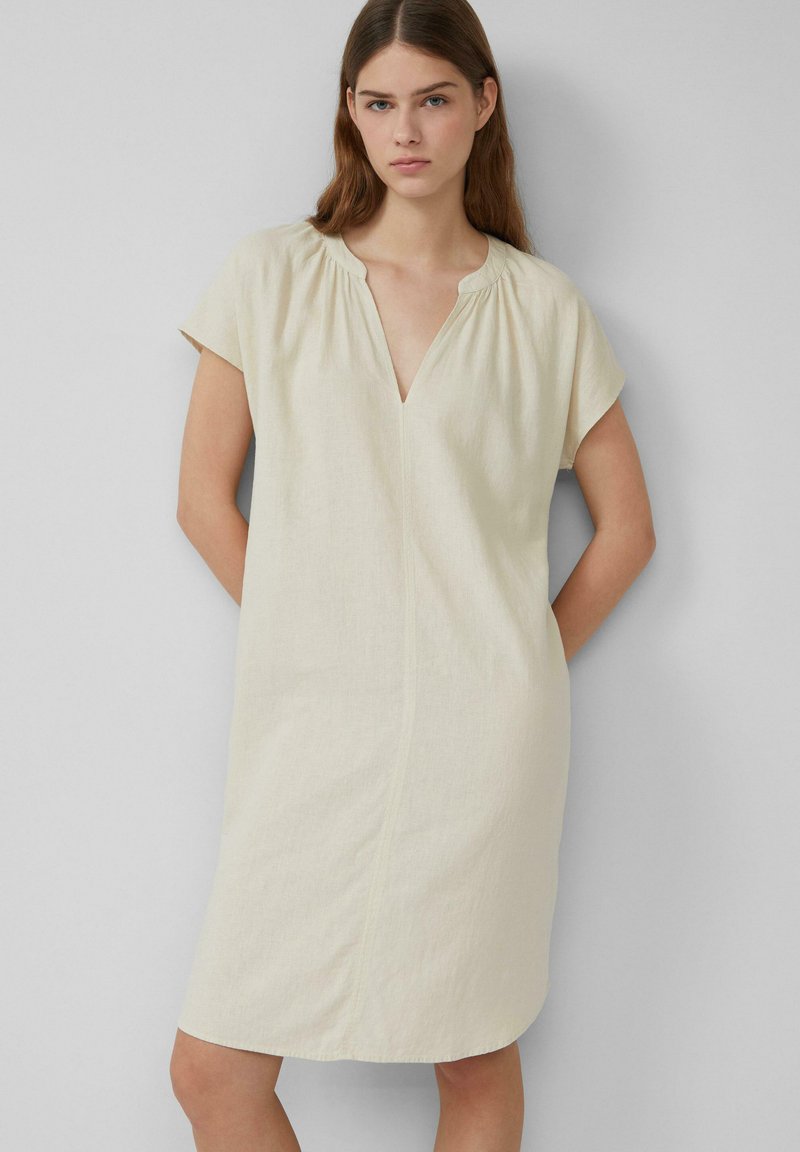 Helles beiges Leinenkleid mit kurzen Ärmeln, V-Ausschnitt und subtiler Raffung am Halsauschnitt. Es hat einen geraden Schnitt und fällt knapp über die Knie.