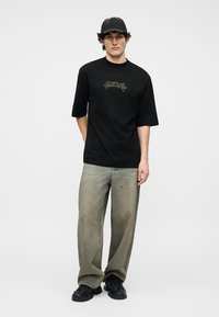 Tânăr care poartă un tricou negru cu imprimeu grafic, blugi largi gri deschiși, decolorați și cu pete, adidași negri și o șapcă neagră de baseball, stând în picioare pe fundal alb.