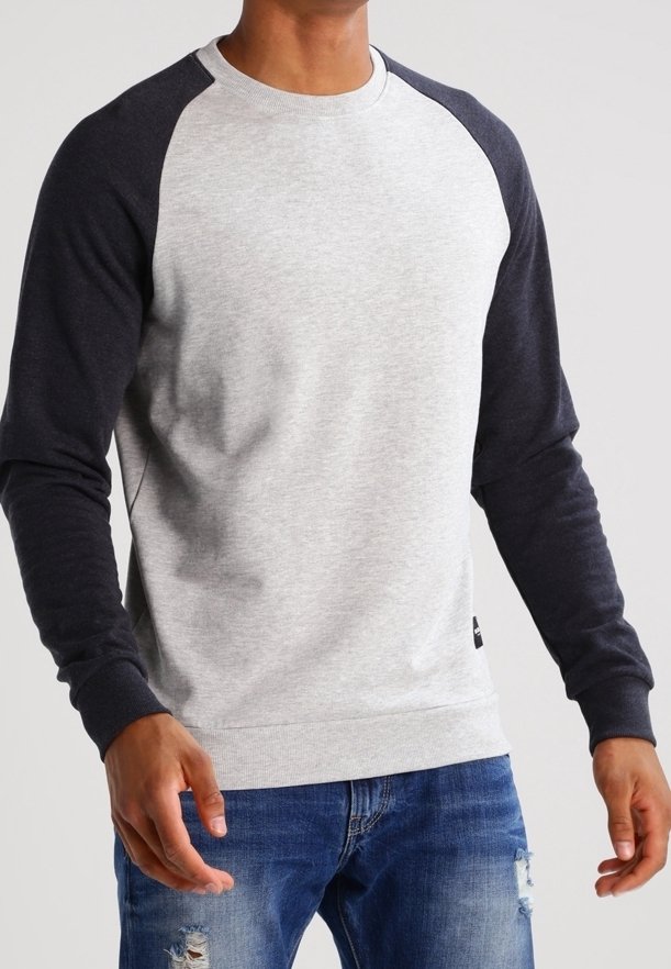 Homme portant un sweat-shirt gris clair avec des manches gris foncé et un jean bleu déchiré, debout devant un fond uni.