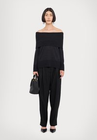 Pull noir à épaules dénudées, manches longues, associé à un pantalon noir à rayures. Le modèle tient un petit sac noir avec des éléments en or.