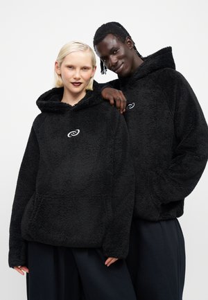 Deux personnes portant des sweats à capuche en polaire noire avec un petit logo blanc sur la poitrine posent devant un fond blanc uni.