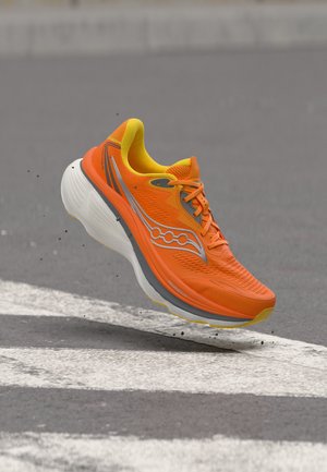 Chaussure de course orange vif avec des accents jaunes, en l'air au-dessus d'une route en asphalte grise avec des marquages blancs, petits débris éparpillés autour.