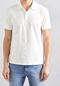 Piké i mjuk, krämfärgad bomull med krage, korta ärmar och en subtil logoprydnad. Skräddarsydd passform med rak nederkant och minimala sömmar.