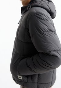 Schwarze Steppjacke mit Kapuze, gestaltet mit einem gesteppten Muster, Frontreißverschluss und Seitentaschen. Weiches, leichtes Material mit glatter Textur.