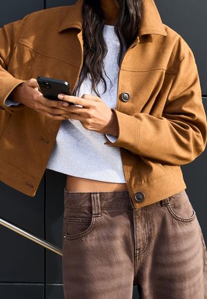 Femme portant une veste et un pantalon marron utilisant un smartphone avec les deux mains devant un mur sombre.