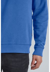 Sudadera azul con puños y dobladillo acanalados, hecha de un tejido suave. Presenta una pequeña etiqueta de marca en el lado inferior. Diseño sencillo y sólido.