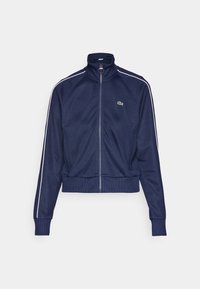 Giacca zip-up blu navy con colletto alto, polsini e fondo a coste, con strisce laterali bianche e un piccolo logo verde sul petto.