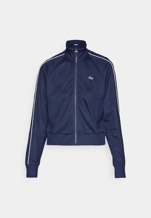Marineblauwe ritsjack met hoge kraag, ribgebreide manchetten en zoom, met witte zijkanten en een klein groen logo op de borst.