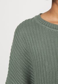 Maglione verde lavorato a maglia con un motivo testurizzato, collo rotondo e polsini a coste. Il tessuto appare morbido e traspirante.