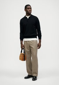 Μαύρο πλεκτό πόλο, εκρού φανελένιο μπλουζάκι, μπεζ μάλλινο παντελόνι και μαύρα παπούτσια. Κρατά μια μικρή καφέ υφασμάτινη τσάντα με λουράκι.