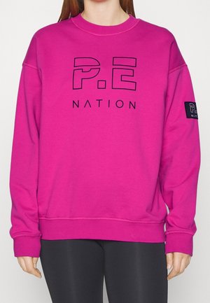 Person iført en knaldrød pink sweatshirt med "P.E Nation"-logo på fronten og ærmet, kombineret med sorte leggings, mod en ensfarvet baggrund.
