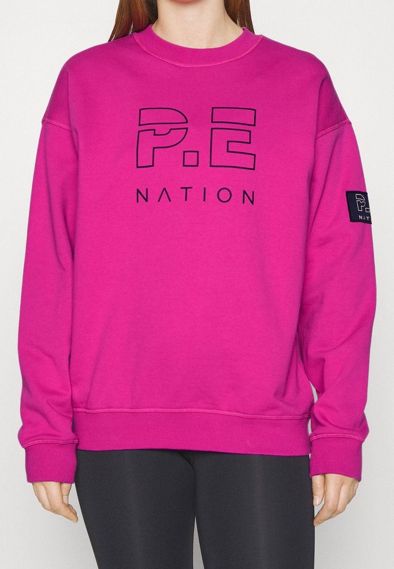 Personne portant un sweat-shirt rose vif avec le logo "P.E Nation" sur le devant et la manche, assorti à un legging noir, sur un fond uni.