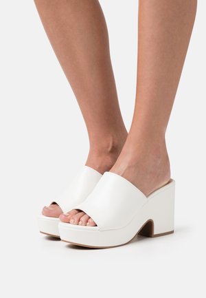 Mules blancs à plateforme avec une tige en cuir lisse, bout ouvert, forme arrondie et un large talon bloc. Présente un design minimaliste.