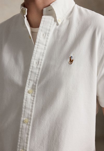 Férfi, fehér gombos ingben, amelynek bal mellrészén egy kis hímzett póljátékos logó látható, nyaktól a mellkas közepéig.