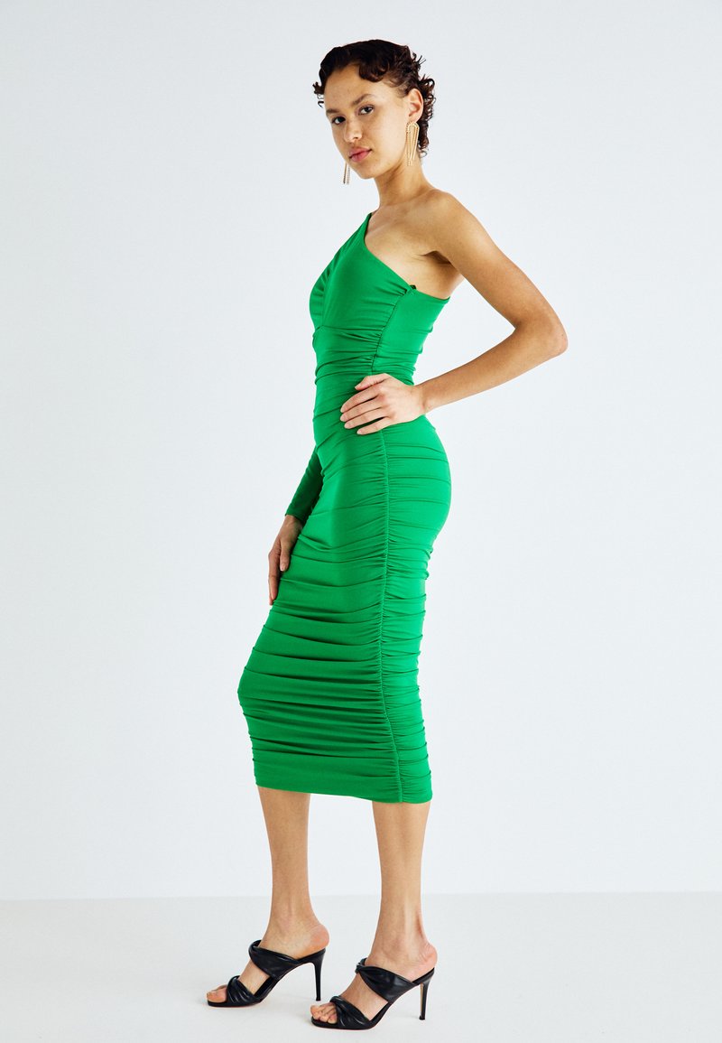 Πράσινο φόρεμα με έναν ώμο, που διαθέτει υφή με ρυτίδες, εφαρμοστή σιλουέτα και μήκος μέχρι το γόνατο. Συνδυάζεται με μαύρες ψηλοτάκουνες σαγιονάρες.