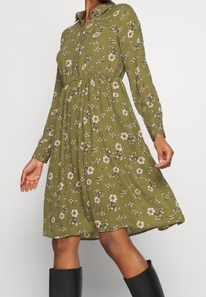 Zielona sukienka w kwiaty z długim rękawem, kołnierzykiem, marszczoną talią i długością do kolan, noszona z czarnymi butami.