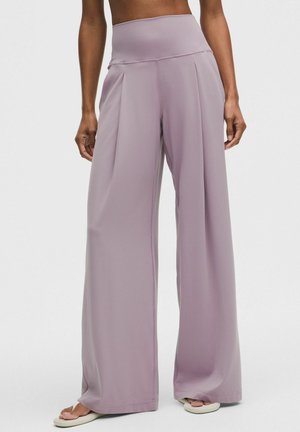 Kvinde iført højtaljede, vidde ben, lavendelfarvede bukser sammen med hvide klip-klappere mod en ensfarvet baggrund.