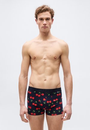 Νεαρός άνδρας με σγουρά μαλλιά που φοράει μαύρα βαμβακερά εσώρουχα με μοτίβο από κόκκινα κεράσια, στέκεται μπροστά σε ένα απλό ανοιχτό γκρι φόντο.