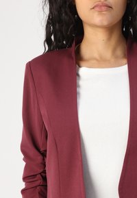Burgundský blazer s hladkou texturou a šálovým límcem, doplněný bílou přiléhavou košilí pod ním. Detailní záběr horní části oděvu.
