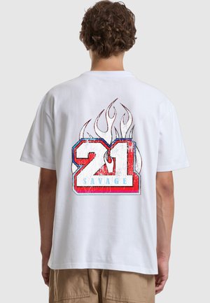 Person, die ein weißes T-Shirt mit einer großen rot-weiß-blauen „21 Savage“-Grafik und Flammendesign auf dem Rücken trägt.