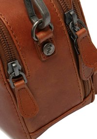 Bolso de cuero marrón con herrajes de metal plateado, que presenta cremalleras, costuras beige y un detalle de logo circular en relieve. Diseño compacto.