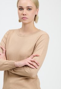 Maglione beige lavorato a maglia con maniche lunghe, scollatura rotonda e dettagli arricciati sui polsini. Texture liscia con una leggera finitura scintillante.