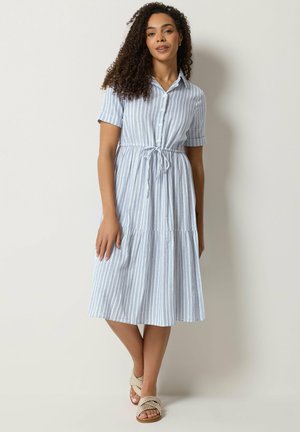 Kobieta z kręconymi włosami, ubrana w błękitno-białą sukienkę midi w paski z krótkim rękawem i paskiem w talii, stoi na tle jednolitej ściany.