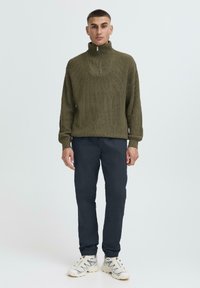 Olive green ribbed sweater with a high collar and half zipper, paired with dark navy trousers and beige athletic shoes.