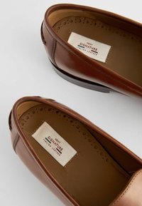 Braune Lederloafers mit glänzender Oberfläche, subtilem Design, runder Zehenform und geprägtem Innenfutter mit durchbrochener Detailgestaltung.