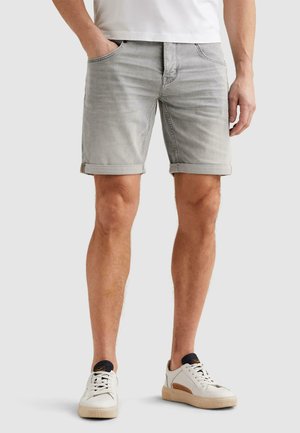 Mand iført lysegrå denimshorts med opsmøgede kanter, hvide sneakers med sort detalje og en hvid skjorte, stående foran en ensfarvet baggrund.