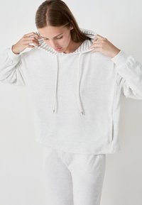 Sudadera corta de color gris claro, hecha de una tela suave, con capucha de interior a rayas y cordones. Combinada con pantalones ligeros a juego.