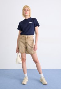 T-shirt en coton navy avec logo, short en jean beige taille haute, chaussettes blanches à la cheville, baskets beiges et sac texturé blanc cassé.
