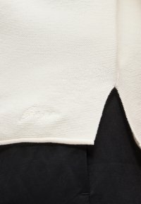 Κοντινή λήψη πλεκτού υφάσματος σε κρεμ χρώμα με διακριτικό υφασμάτινο μοτίβο και μικρή πλαϊνή σχισμή πάνω σε μαύρο φόντο υφάσματος.