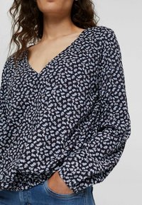 Donna che indossa un blusa nera con un delicato motivo floreale blu e bianco e maniche lunghe, abbinata a jeans blu, con la mano nella tasca.