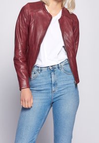 Giubbotto in pelle bordeaux con colletto a costine, chiusura con zip e design aderente, indossato sopra una t-shirt bianca e jeans azzurri chiari.