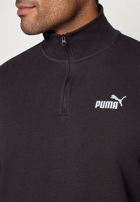 Sweatshirt noir à col montant avec une fermeture éclair quart. Présente un petit logo Puma blanc sur la poitrine.