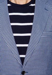 Niebieska i biała marynarka w wzór houndstooth z jednym czarnym guzikiem, noszona na niebieskiej i białej koszuli w paski. Teksturowany materiał z wyraźnymi klapami.