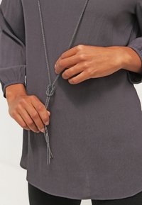 Graue Tunika mit rundem Ausschnitt und langen Ärmeln. Eine silberne Kettenhalskette mit Knoten-Akzent und Quasten. Hände halten die Halskette.